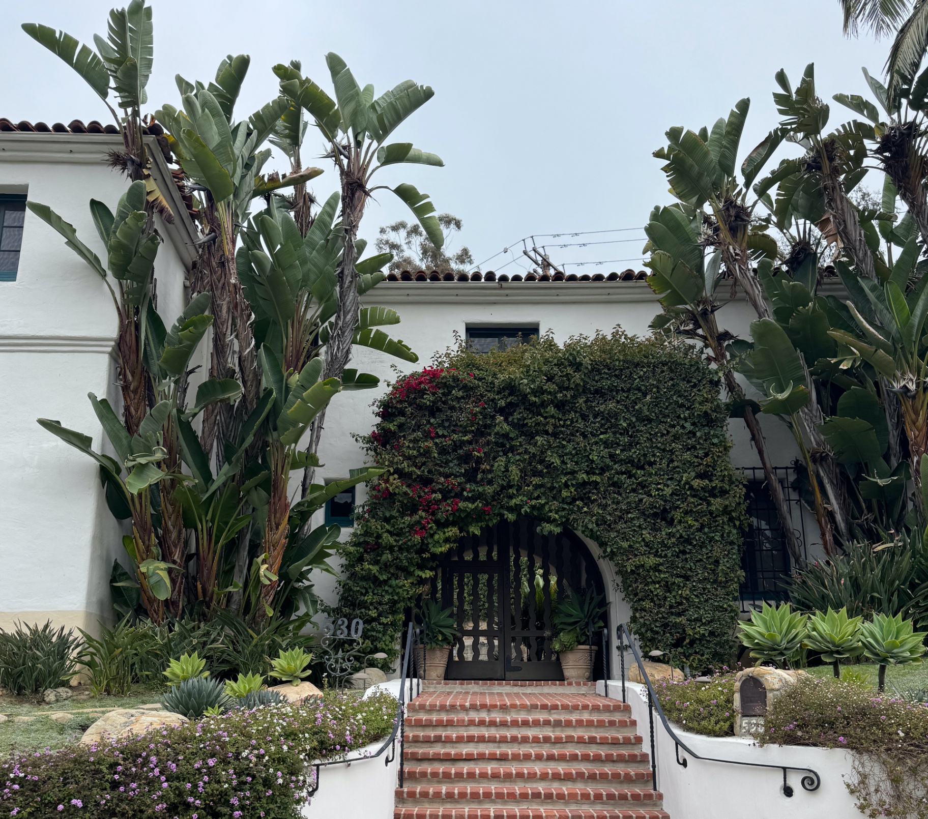 Beautiful Spanish Colonial Revival home in Camarillo CA with white stucco walls, red clay tile roof, lush tropical landscaping featuring banana plants and bougainvillea, brick walkway and arched entrance - professional house cleaning services for luxury homes, family residences, and suburban communities throughout Camarillo including Sterling Hills, Mission Oaks, Spanish Hills, and Premium Outlets area